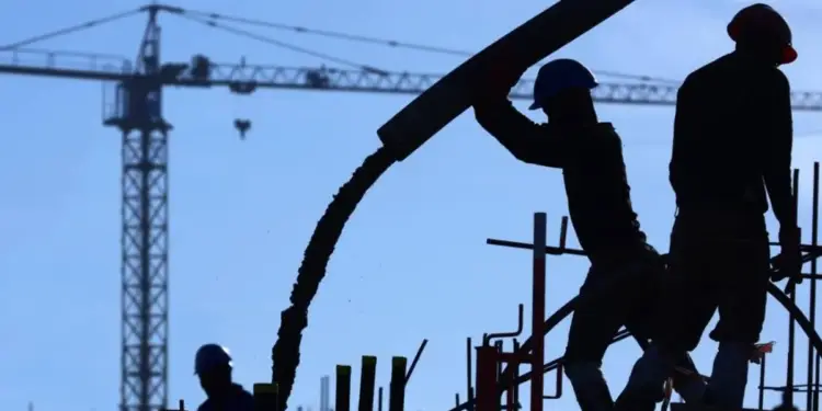 Siluetas de trabajadores de construcción vertiendo concreto con una bomba en una obra. Se observa una grúa torre y andamios bajo un cielo azul claro.
