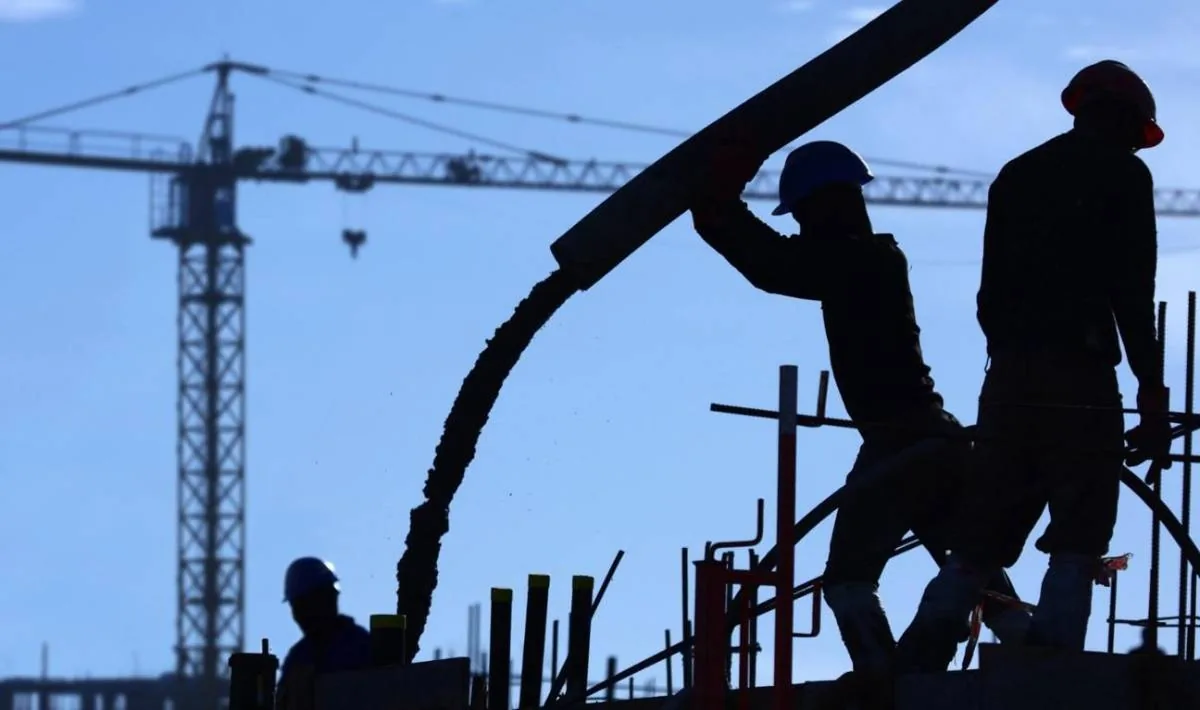 Siluetas de trabajadores de construcción vertiendo concreto con una bomba en una obra. Se observa una grúa torre y andamios bajo un cielo azul claro.
