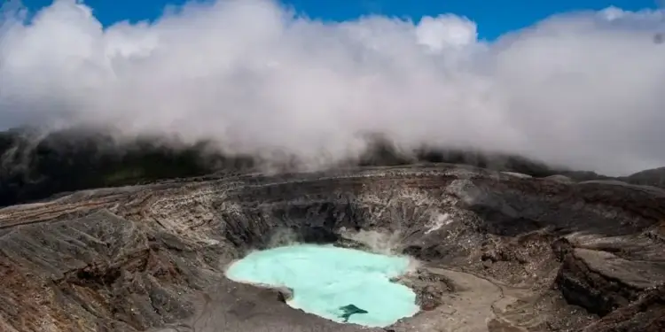 Vista panorámica de un cráter volcánico profundo que contiene una laguna de aguas color turquesa rodeada de rocas y nubes.