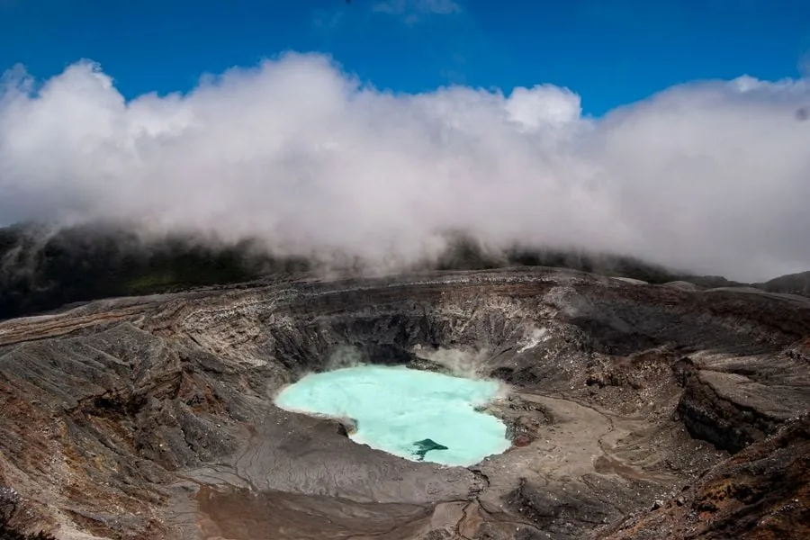 Vista panorámica de un cráter volcánico profundo que contiene una laguna de aguas color turquesa rodeada de rocas y nubes.
