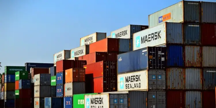 Gran pila de contenedores de carga de varios colores (rojo, azul, verde, blanco) apilados en un puerto o terminal logística bajo un cielo azul claro. Algunos muestran marcas como Maersk y Evergreen.