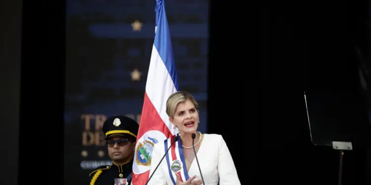 Mujer con banda presidencial hablando frente a un atril con micrófono y una bandera nacional al fondo.