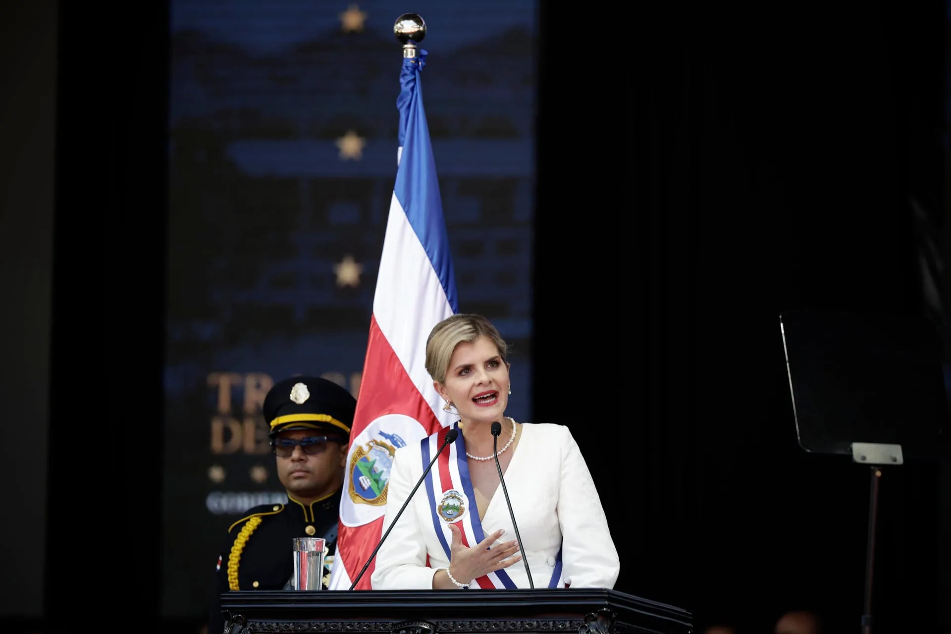 Mujer con banda presidencial hablando frente a un atril con micrófono y una bandera nacional al fondo.