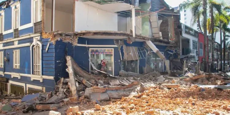 Fachada de un edificio de dos pisos pintado de azul con daños estructurales severos y escombros en la calle.