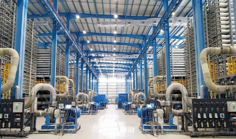 Vista interior de una gran planta industrial con estructuras metálicas azules y complejos sistemas de tuberías y maquinaria.