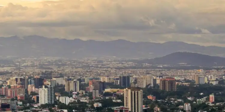 Vista aérea de una ciudad con edificios modernos y residenciales, rodeada de vegetación y montañas, bajo un cielo nublado al atardecer.