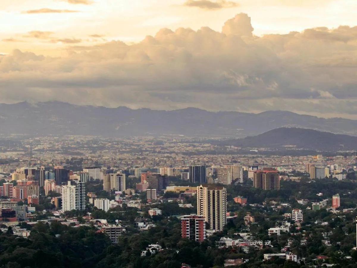 Vista aérea de una ciudad con edificios modernos y residenciales, rodeada de vegetación y montañas, bajo un cielo nublado al atardecer.