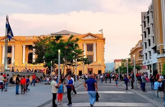 Personas caminando en una amplia plaza pública frente a un gran edificio histórico de color amarillo.