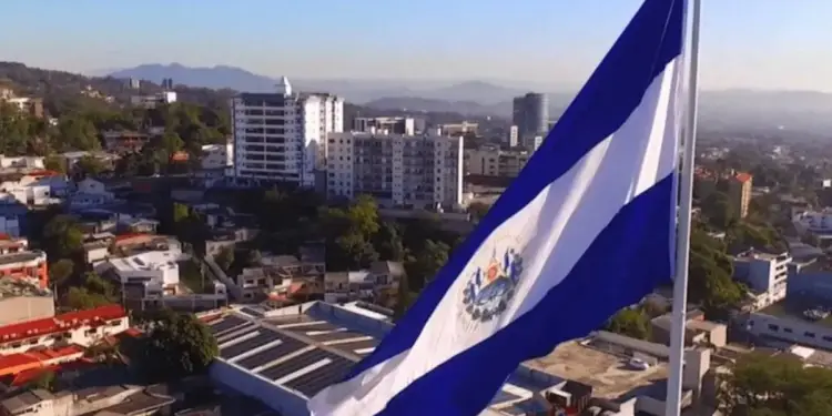 Vista aérea de la bandera de El Salvador ondeando sobre un paisaje urbano con edificios modernos, casas y montañas al fondo. Se aprecian paneles solares en un techo.