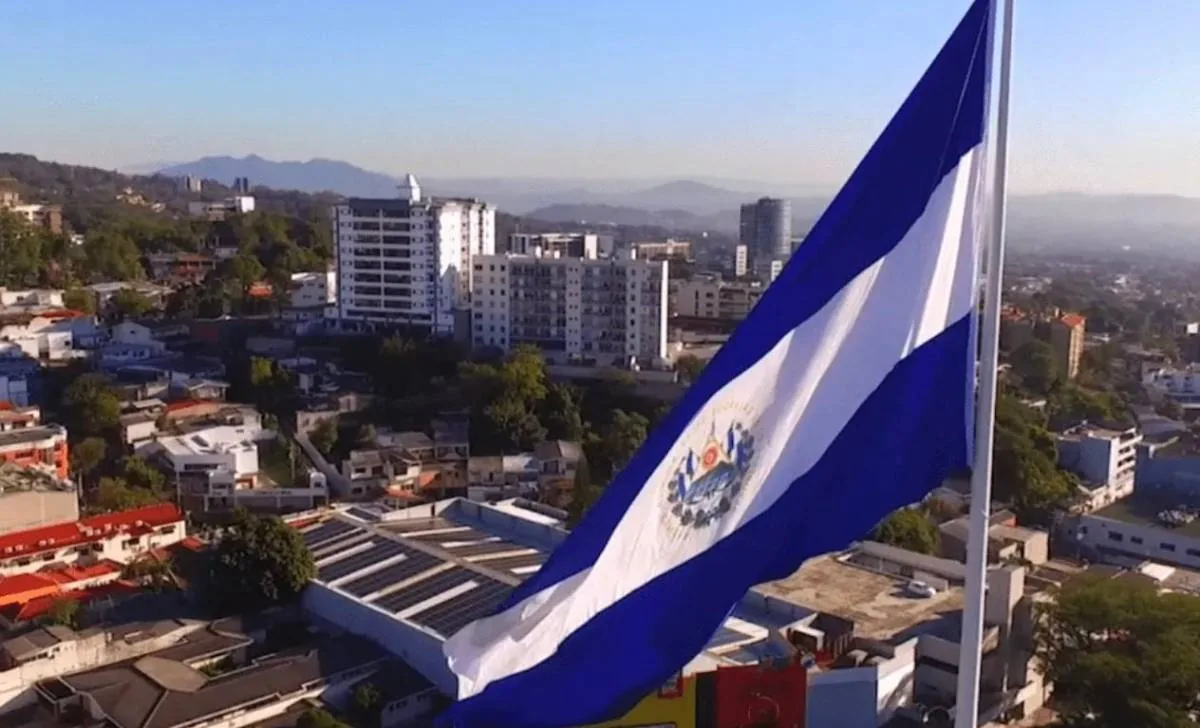 Vista aérea de la bandera de El Salvador ondeando sobre un paisaje urbano con edificios modernos, casas y montañas al fondo. Se aprecian paneles solares en un techo.
