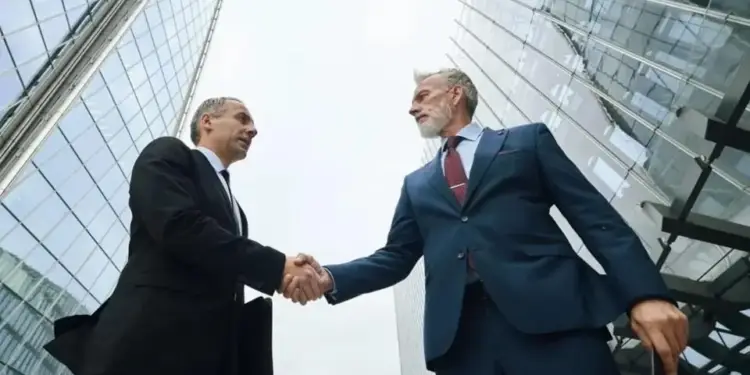 Dos ejecutivos en traje se estrechan la mano con edificios modernos de cristal de fondo, representando un acuerdo profesional.