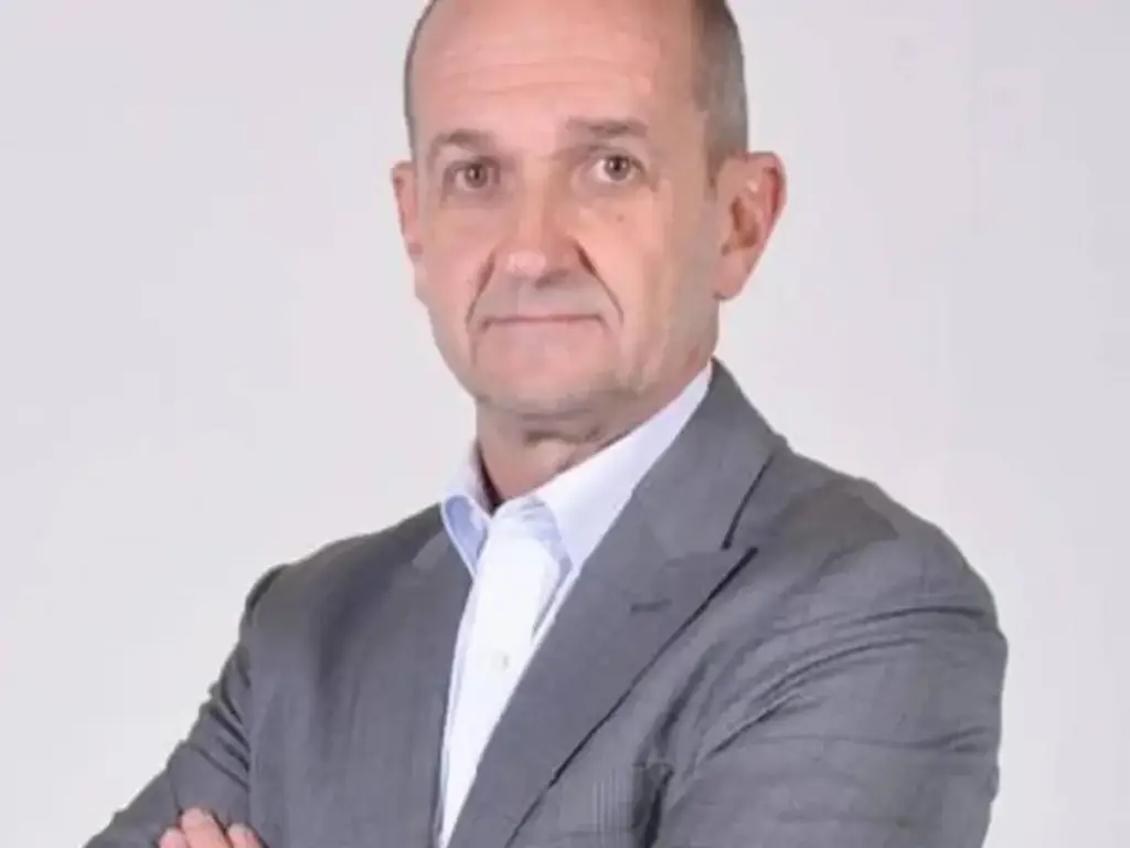 Retrato de un hombre de mediana edad, calvo, con traje gris y camisa azul claro, brazos cruzados, mirando al frente con expresión seria sobre fondo blanco.