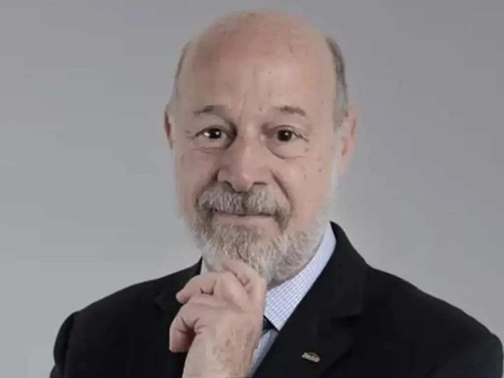 Retrato de un hombre mayor calvo con barba gris, vestido con traje oscuro y camisa a cuadros, con la mano en la barbilla, mirando pensativo.