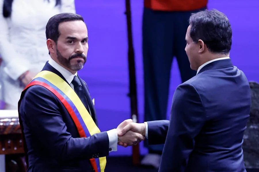 Dos hombres con traje formal estrechándose la mano en un evento oficial con fondo morado.