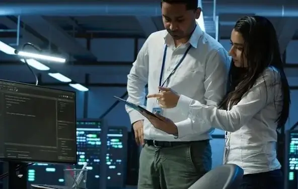 Dos profesionales analizando datos en una tablet dentro de una sala de servidores con monitores iluminados.