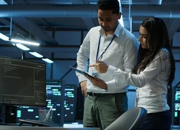 Dos profesionales analizando datos en una tablet dentro de una sala de servidores con monitores iluminados.