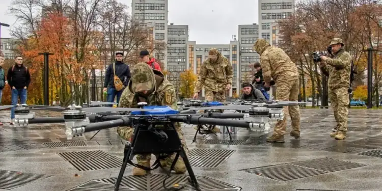 Un grupo de personas, algunas en uniforme, examina drones profesionales en una plaza urbana. Edificios y árboles de otoño al fondo.