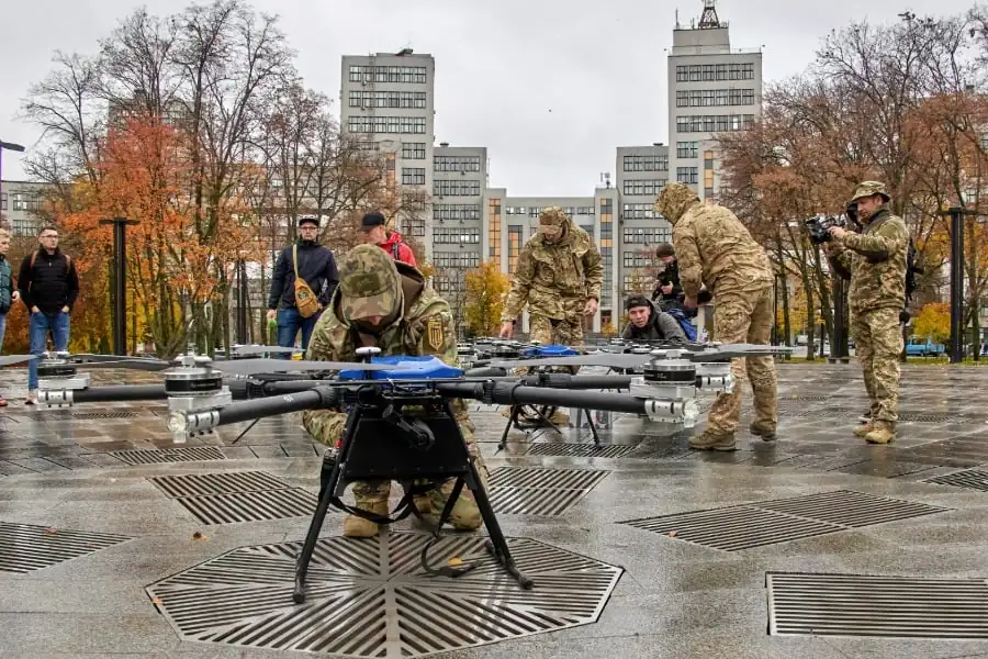 Un grupo de personas, algunas en uniforme, examina drones profesionales en una plaza urbana. Edificios y árboles de otoño al fondo.