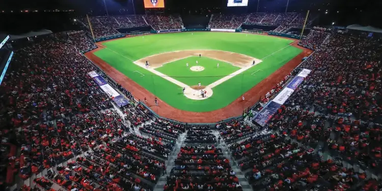 Vista aérea nocturna de un estadio de béisbol repleto de aficionados. Campo verde iluminado con jugadores y pantallas gigantes.
