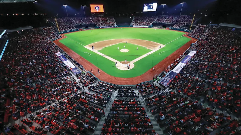 Vista aérea nocturna de un estadio de béisbol repleto de aficionados. Campo verde iluminado con jugadores y pantallas gigantes.