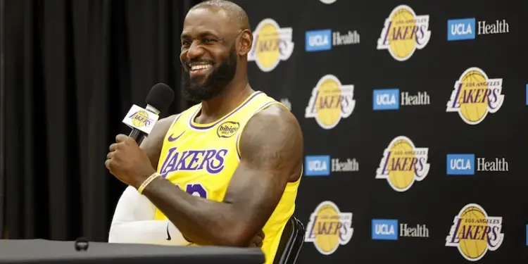 Hombre sonriente con barba, vistiendo camiseta amarilla de baloncesto con 'LAKERS', sosteniendo un micrófono. Fondo con logos de 'LAKERS' y 'UCLA Health'.