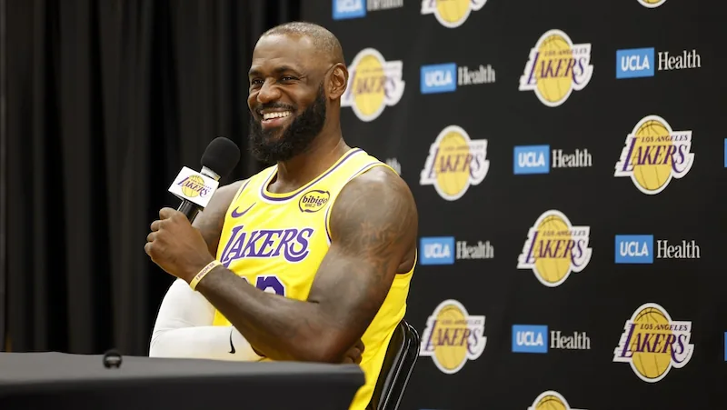 Hombre sonriente con barba, vistiendo camiseta amarilla de baloncesto con 'LAKERS', sosteniendo un micrófono. Fondo con logos de 'LAKERS' y 'UCLA Health'.