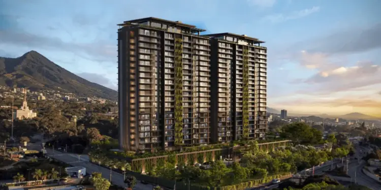 Vista aérea de un moderno edificio de gran altura con fachadas verdes en un entorno urbano montañoso al atardecer, rodeado de vegetación y carreteras.