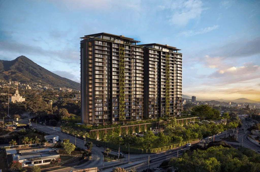 Vista aérea de un moderno edificio de gran altura con fachadas verdes en un entorno urbano montañoso al atardecer, rodeado de vegetación y carreteras.