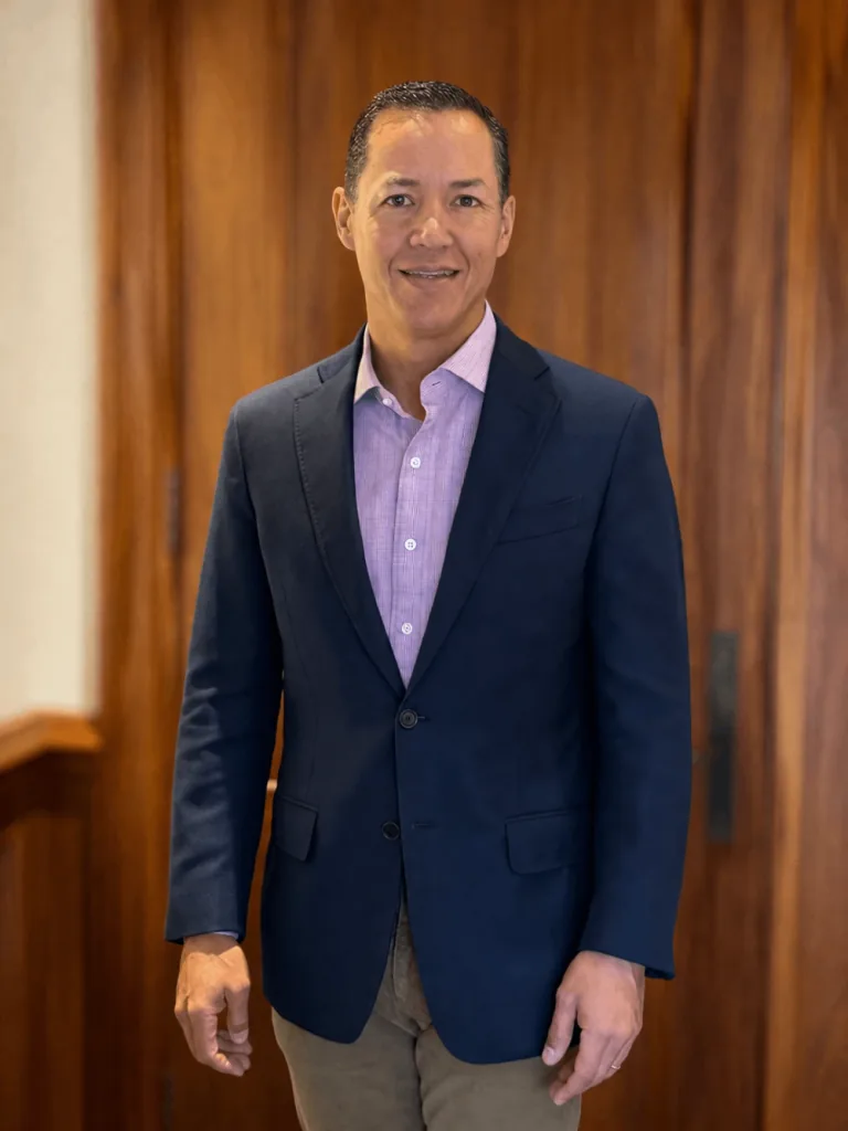 Hombre de mediana edad, tez morena, sonriendo, vestido con un blazer azul marino, camisa lila y pantalón caqui, de pie frente a una pared de madera.
