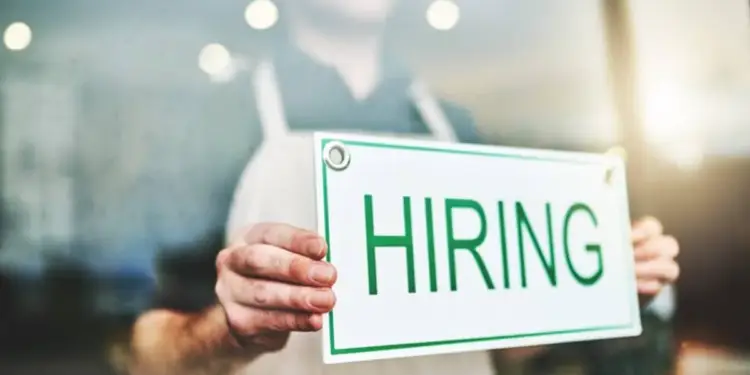 Persona con delantal sosteniendo un cartel blanco con la palabra 'HIRING' en letras verdes. Representa oportunidades de empleo y contratación en un negocio.