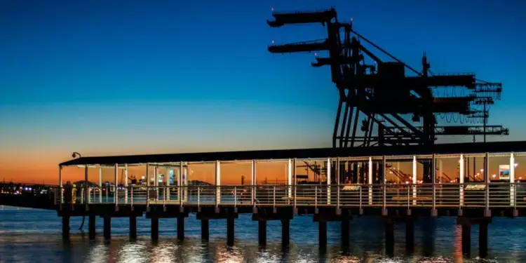 Silueta de grúas portuarias y un muelle moderno iluminado sobre el agua al atardecer, reflejando la actividad comercial y logística marítima.