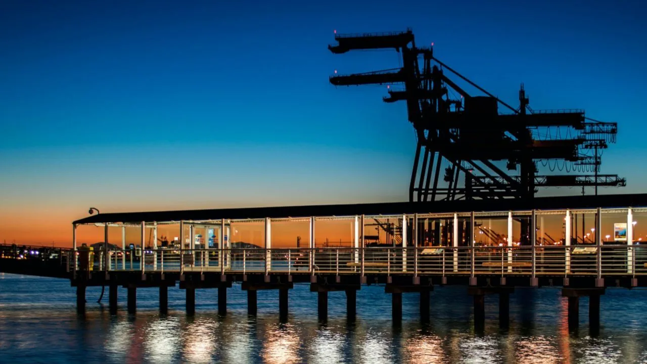Silueta de grúas portuarias y un muelle moderno iluminado sobre el agua al atardecer, reflejando la actividad comercial y logística marítima.