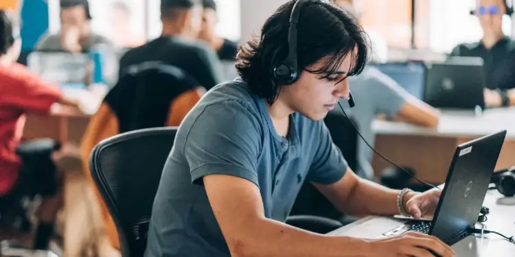 Joven trabajando con ordenador portátil y auriculares con micrófono en una oficina compartida con otros profesionales.