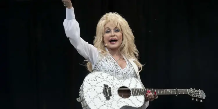 Mujer rubia con cabello voluminoso, vistiendo traje blanco brillante y sosteniendo una guitarra blanca decorada mientras saluda.