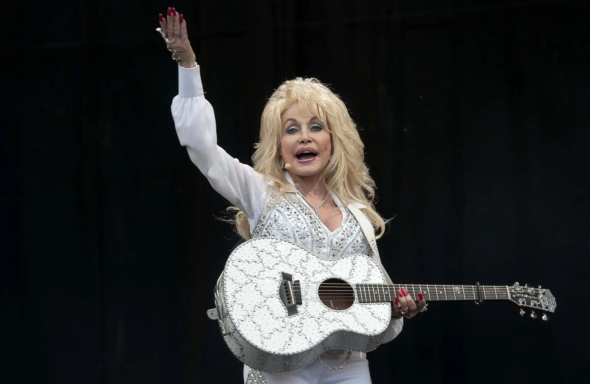 Mujer rubia con cabello voluminoso, vistiendo traje blanco brillante y sosteniendo una guitarra blanca decorada mientras saluda.