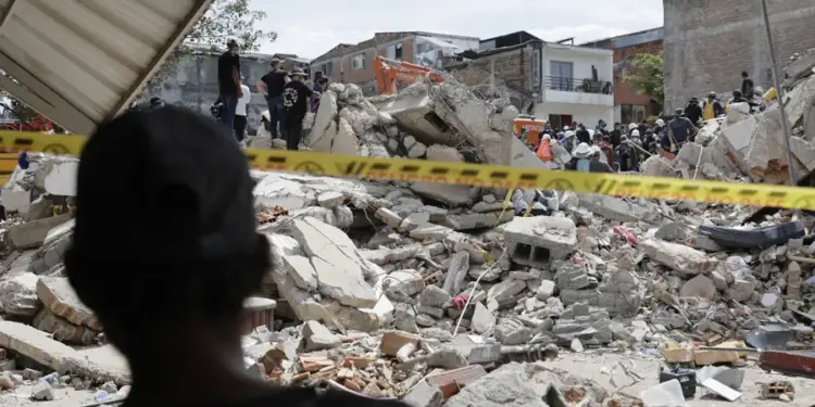 Vista de escombros de edificios colapsados con equipos de rescate y maquinaria pesada, tras una catástrofe. Cinta de precaución amarilla en primer plano.