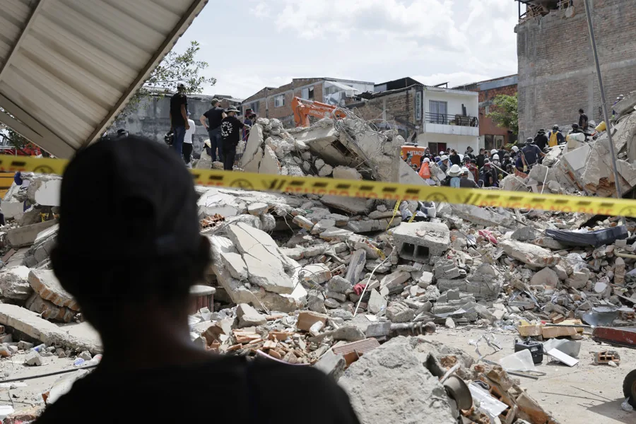 Vista de escombros de edificios colapsados con equipos de rescate y maquinaria pesada, tras una catástrofe. Cinta de precaución amarilla en primer plano.
