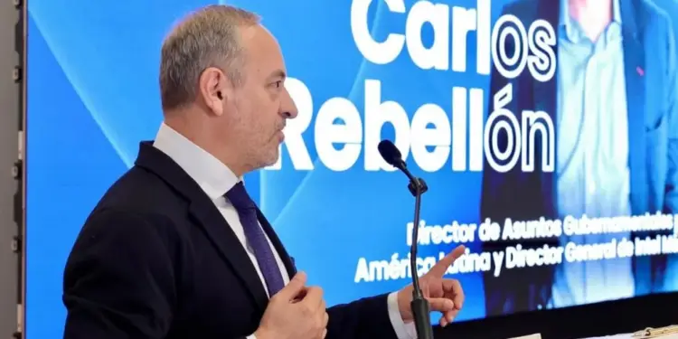 Hombre en traje y corbata hablando en un podio con micrófono. Detrás, una pantalla azul muestra 'Carlos Rebellón' y sus títulos.