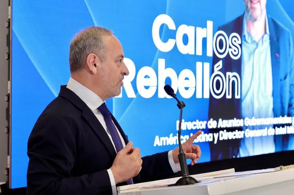 Hombre en traje y corbata hablando en un podio con micrófono. Detrás, una pantalla azul muestra 'Carlos Rebellón' y sus títulos.