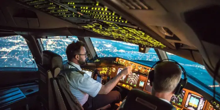 Dos pilotos trabajando en la cabina de mando de un avión comercial durante un vuelo nocturno sobre una ciudad iluminada.