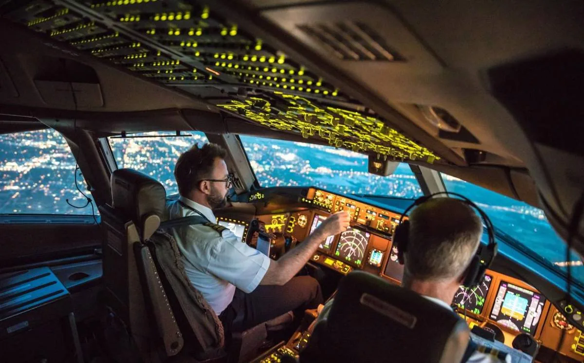 Dos pilotos trabajando en la cabina de mando de un avión comercial durante un vuelo nocturno sobre una ciudad iluminada.