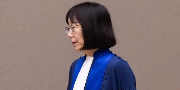 Perfil de mujer con cabello negro y gafas vistiendo una toga azul y blanca contra una pared gris.