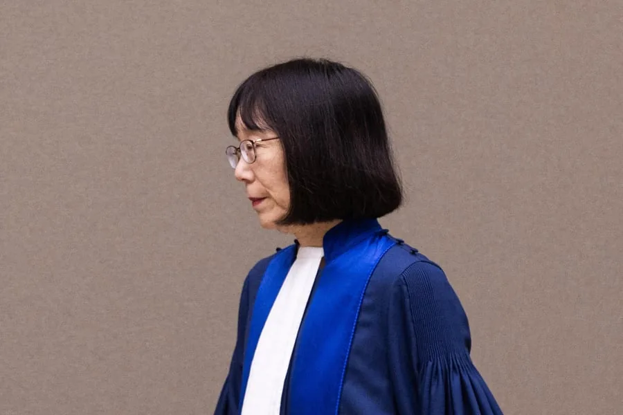 Perfil de mujer con cabello negro y gafas vistiendo una toga azul y blanca contra una pared gris.