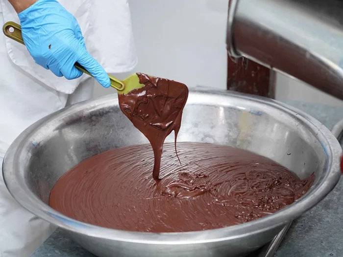 Mano enguantada mezclando chocolate derretido en un gran bol metálico con una espátula verde. Proceso de elaboración de confitería.