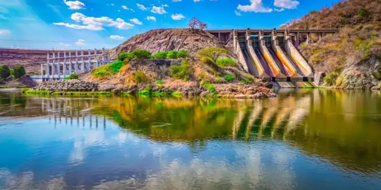 Vista panorámica de una gran presa hidroeléctrica con múltiples compuertas, rodeada de colinas verdes y un río ancho bajo un cielo azul con nubes.