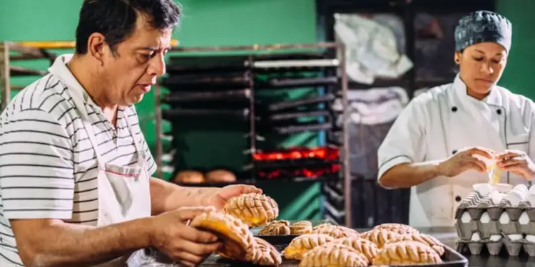 Dos panaderos en una panadería. Un hombre coloca conchas en bandejas, mientras una mujer en uniforme de chef rompe huevos para la preparación.