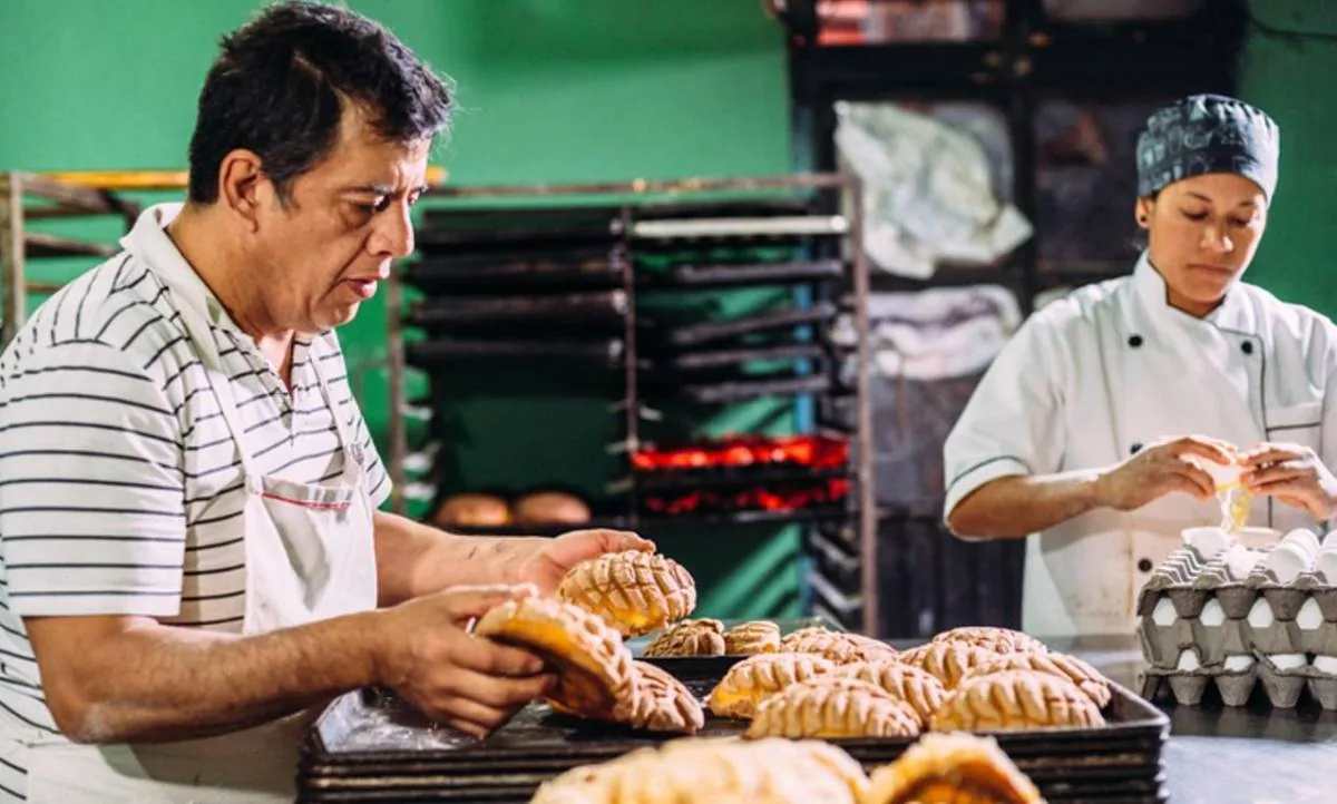 Dos panaderos en una panadería. Un hombre coloca conchas en bandejas, mientras una mujer en uniforme de chef rompe huevos para la preparación.