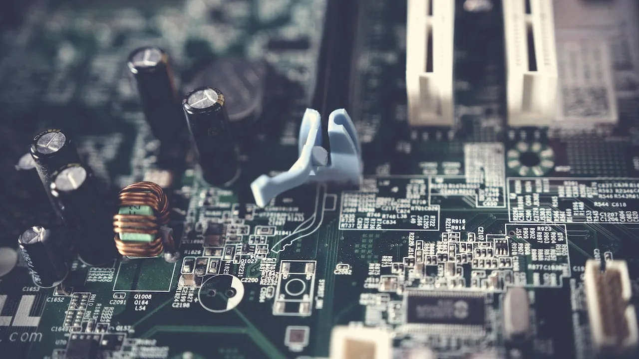 Primer plano de una placa de circuito verde con múltiples componentes electrónicos como condensadores negros, una bobina de cobre y conectores blancos y azules. Representa la tecnología digital.