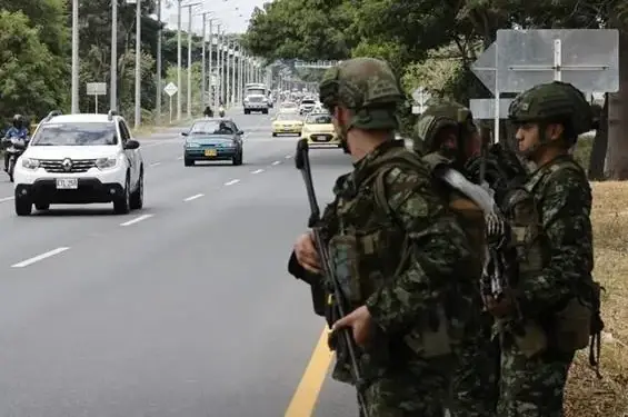 Tres militares armados con uniformes de camuflaje y cascos en el arcén de una carretera con tráfico de vehículos y taxis.