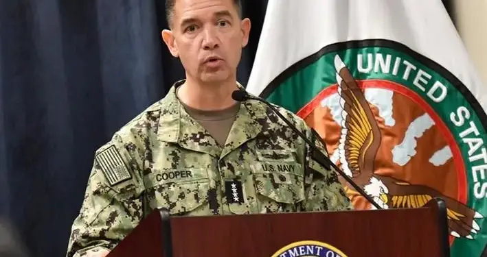 Oficial de la Marina de EE. UU. con uniforme de camuflaje y placa "COOPER" hablando en un podio. Al fondo, una bandera con el emblema de "United States".